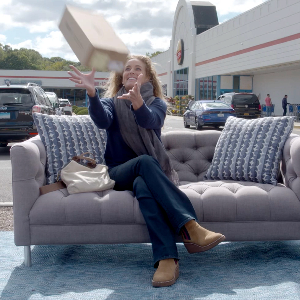 Woman sitting on a couch with a shopping mall behind her catching a box of shoes.